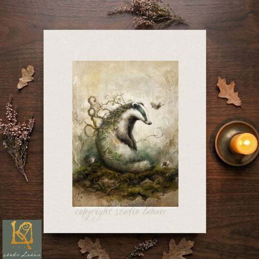 The image is a framed artwork depicting a badger surrounded by a lush, verdant landscape with a bird in flight. The artwork is placed on a wooden surface, with a lit candle and some leaves nearby.