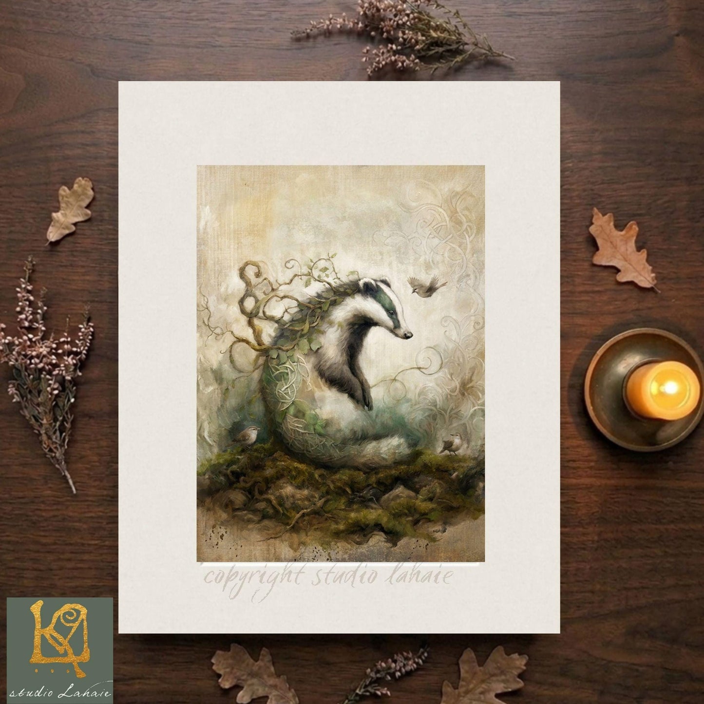 The image is a framed artwork depicting a badger surrounded by a lush, verdant landscape with a bird in flight. The artwork is placed on a wooden surface, with a lit candle and some leaves nearby.